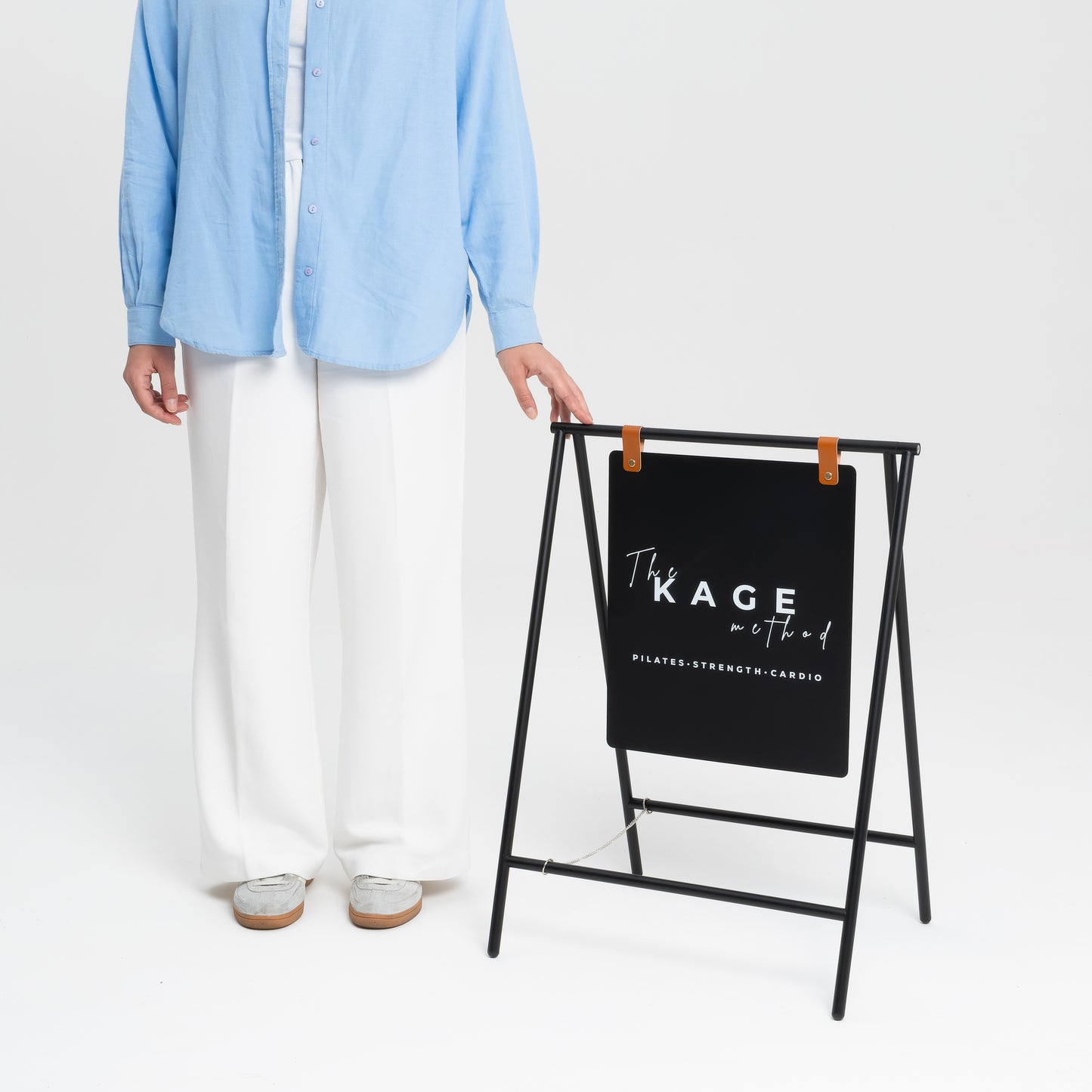 A person standing next to a compact A-frame sidewalk sign with a faux leather strap and text space for customization, displayed in a black color.