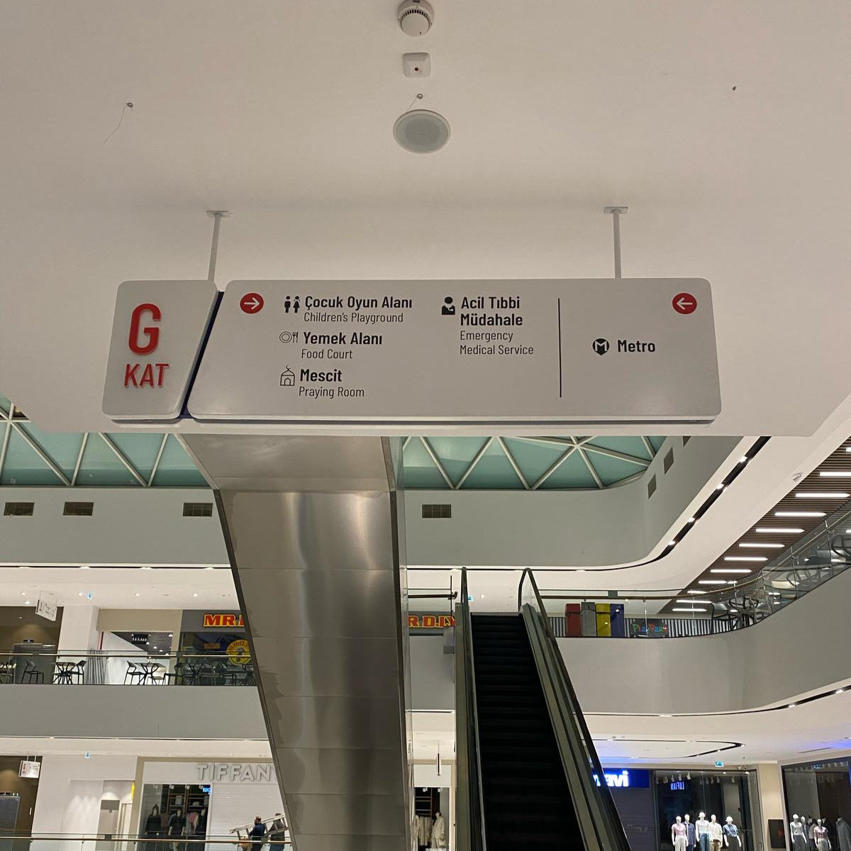 Escalator in a modern shopping mall with a sign indicating 'G Kat' and directions.