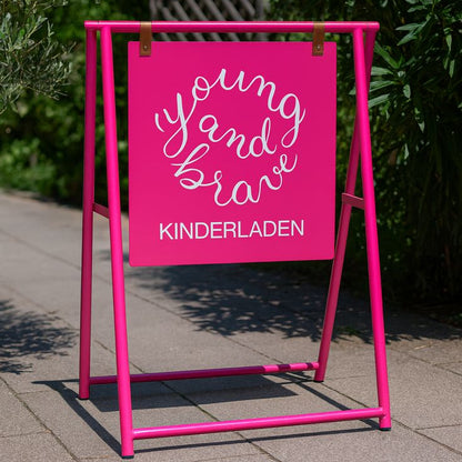 Pink a-frame sign with 'young and brave KINDERLADEN' text on a sidewalk.