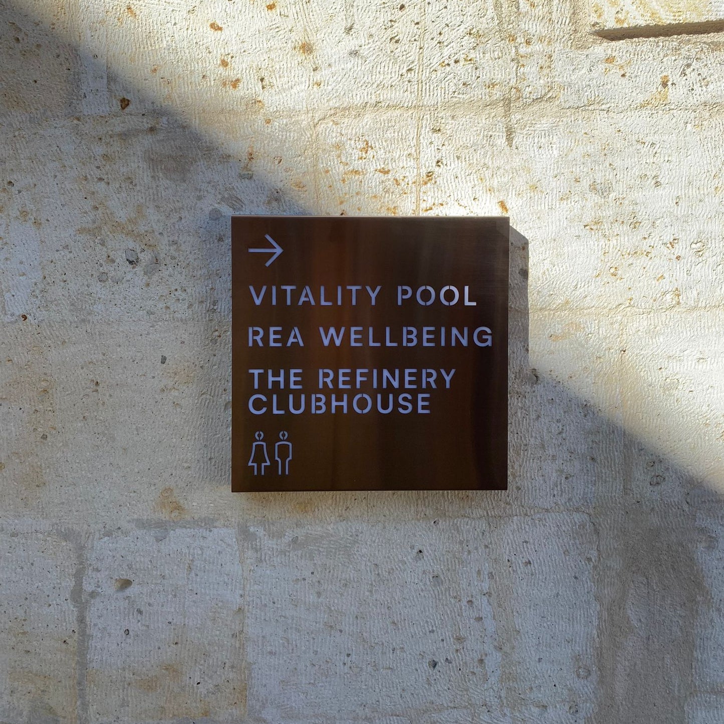 Sign pointing to 'Vitality Pool', 'REA Wellbeing', and 'The Refinery Clubhouse' on a stone wall.