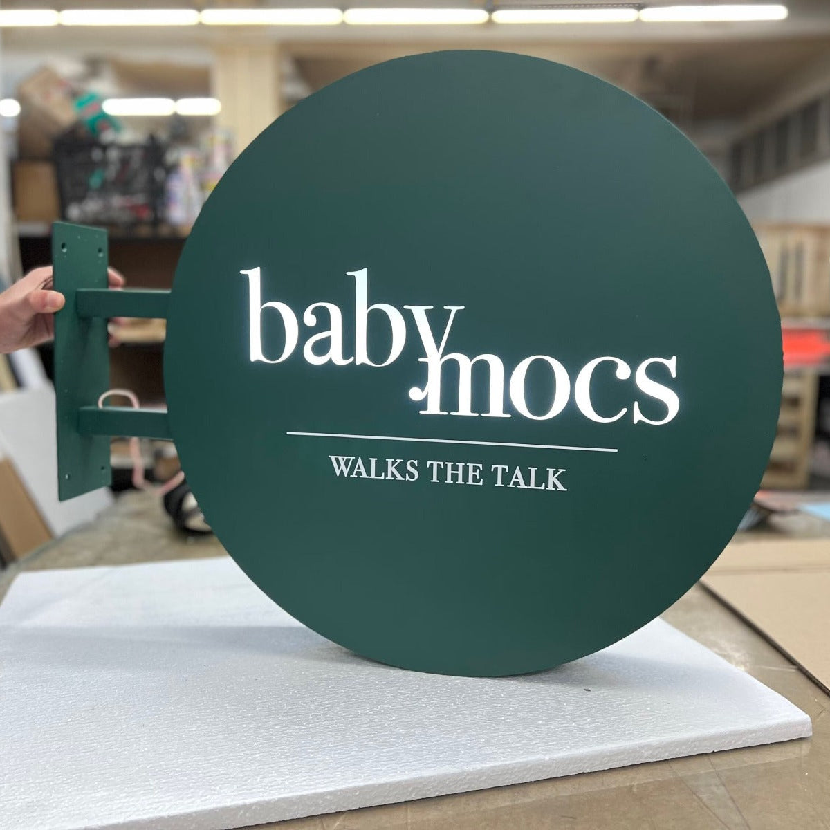 Green circular sign with 'baby mocs' text on a stand in an indoor setting