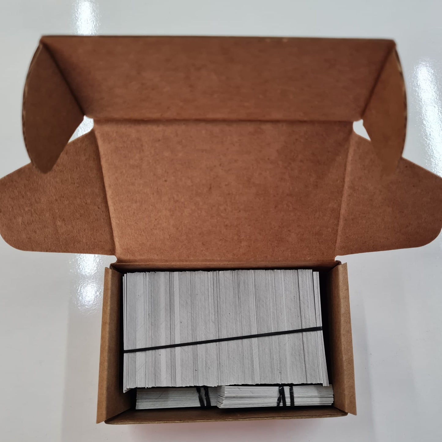 Open brown cardboard box containing a stack of white cards on a white background