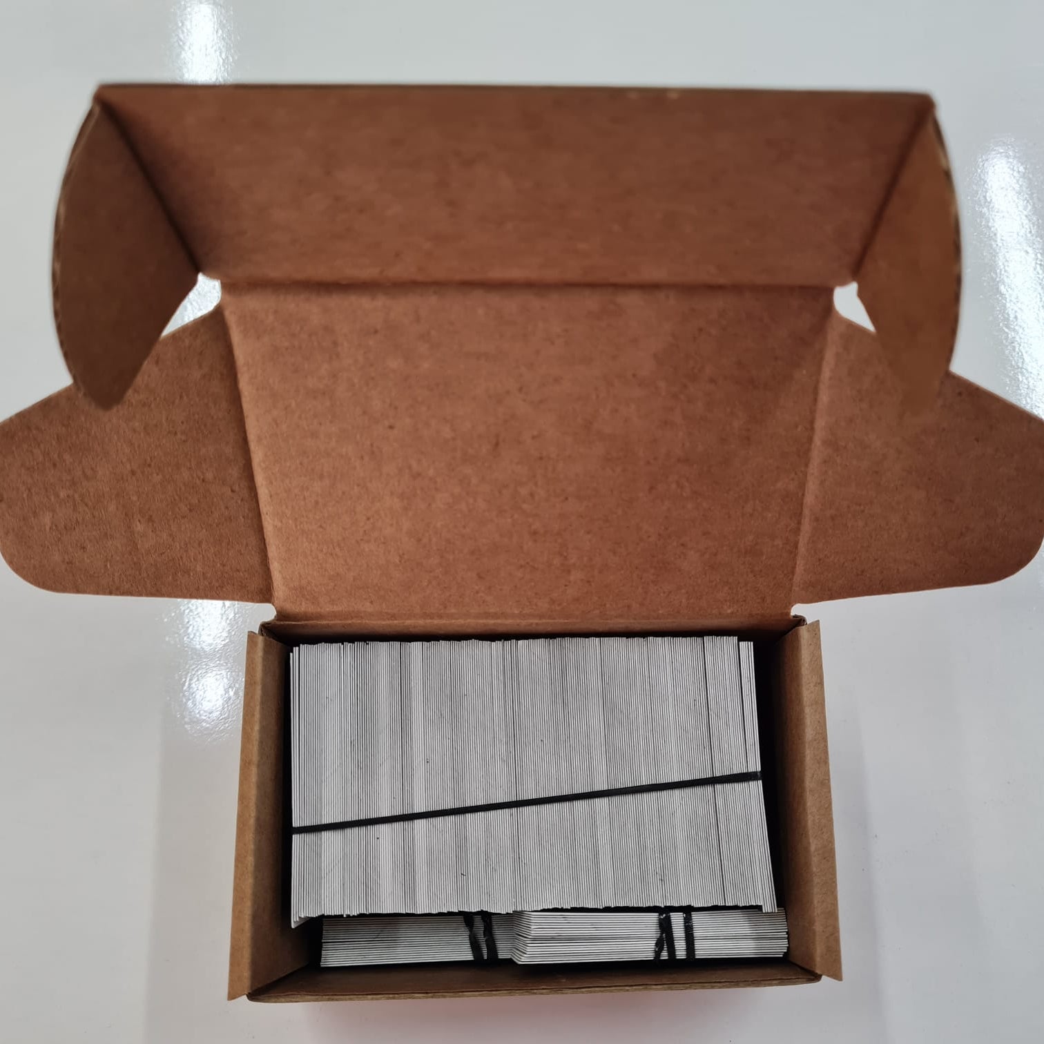 Open brown cardboard box containing a stack of white cards on a white background