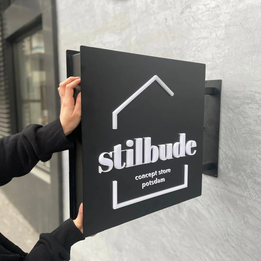 Person adjusting a black sign with 'stilbude' branding on a textured wall.