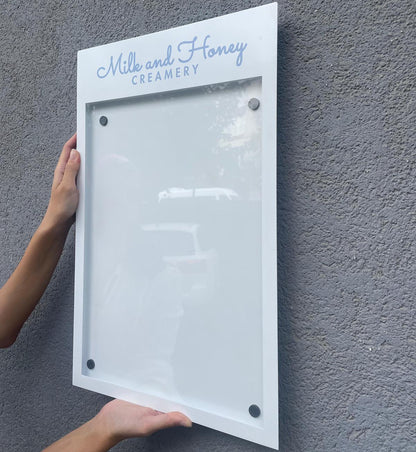 Person mounting a framed sign on a gray wall with 'Milk and Honey Creamery' text.