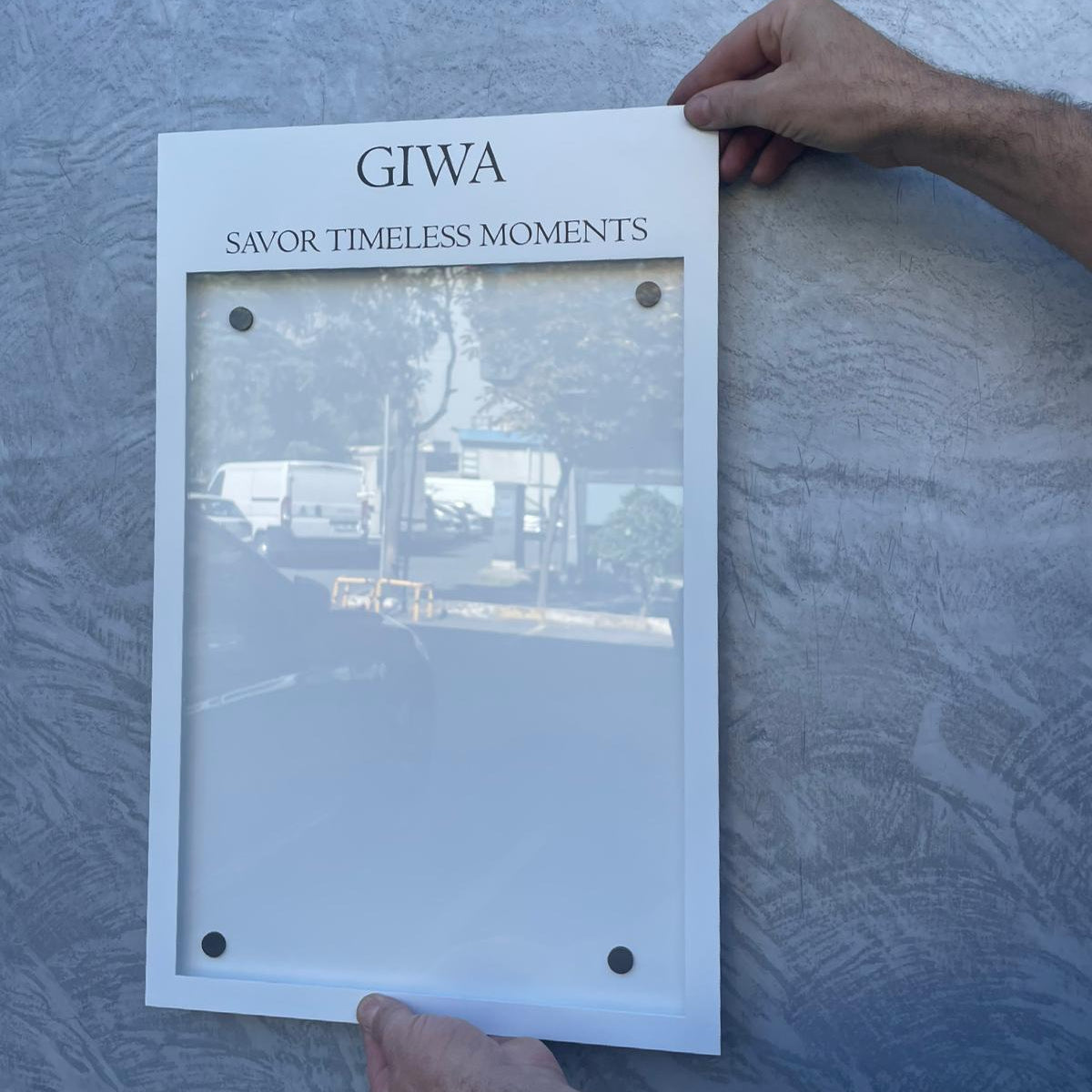 Person holding a white frame with 'GIWA' branding against a textured gray wall.