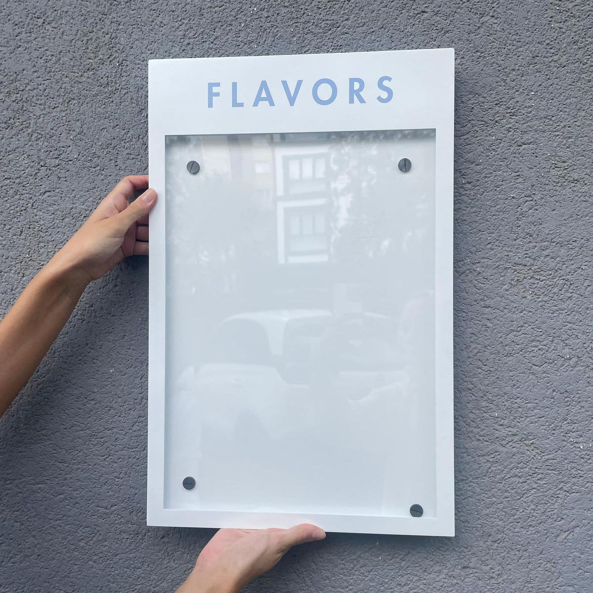 Person holding a white frame with 'FLAVORS' text against a gray wall