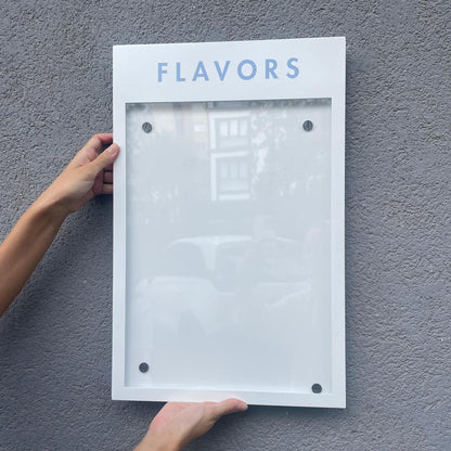 Person holding a white frame with 'FLAVORS' text against a gray wall