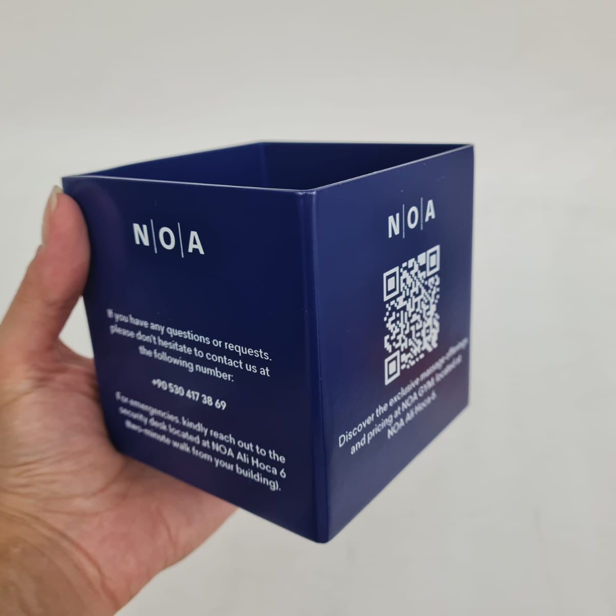 Blue cube-shaped product with 'NOA' branding held by a hand against a gray background
