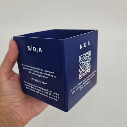 Blue cube-shaped product with 'NOA' branding held by a hand against a gray background