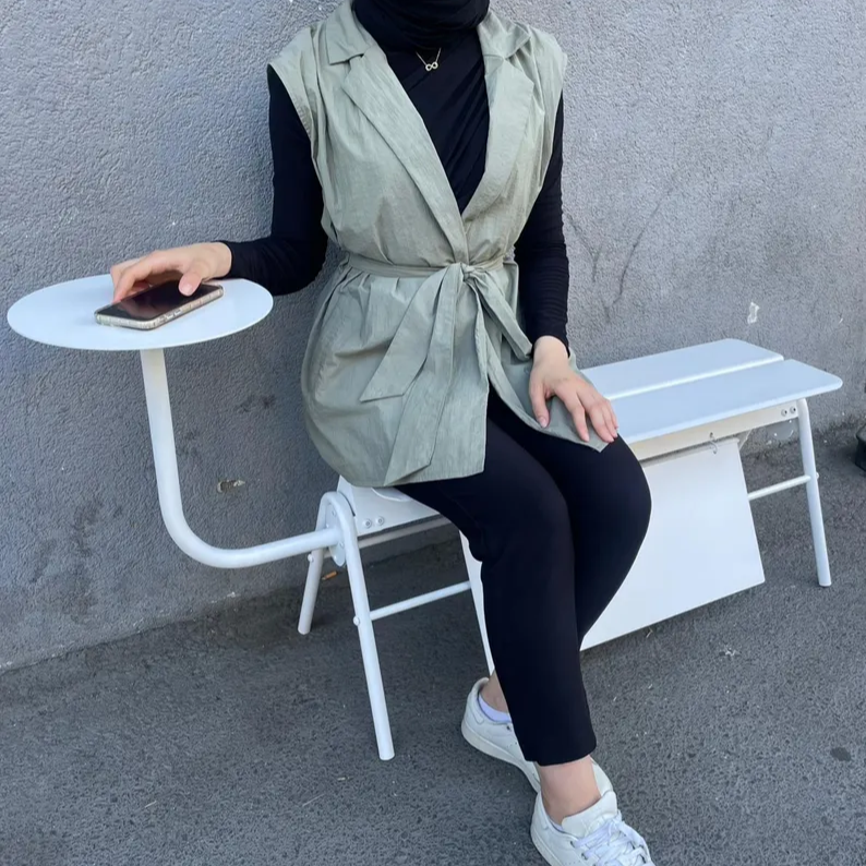 Person sitting on a white bench wearing a green vest and black pants, holding a phone.