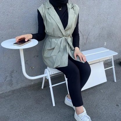 Person sitting on a white bench wearing a green vest and black pants, holding a phone.