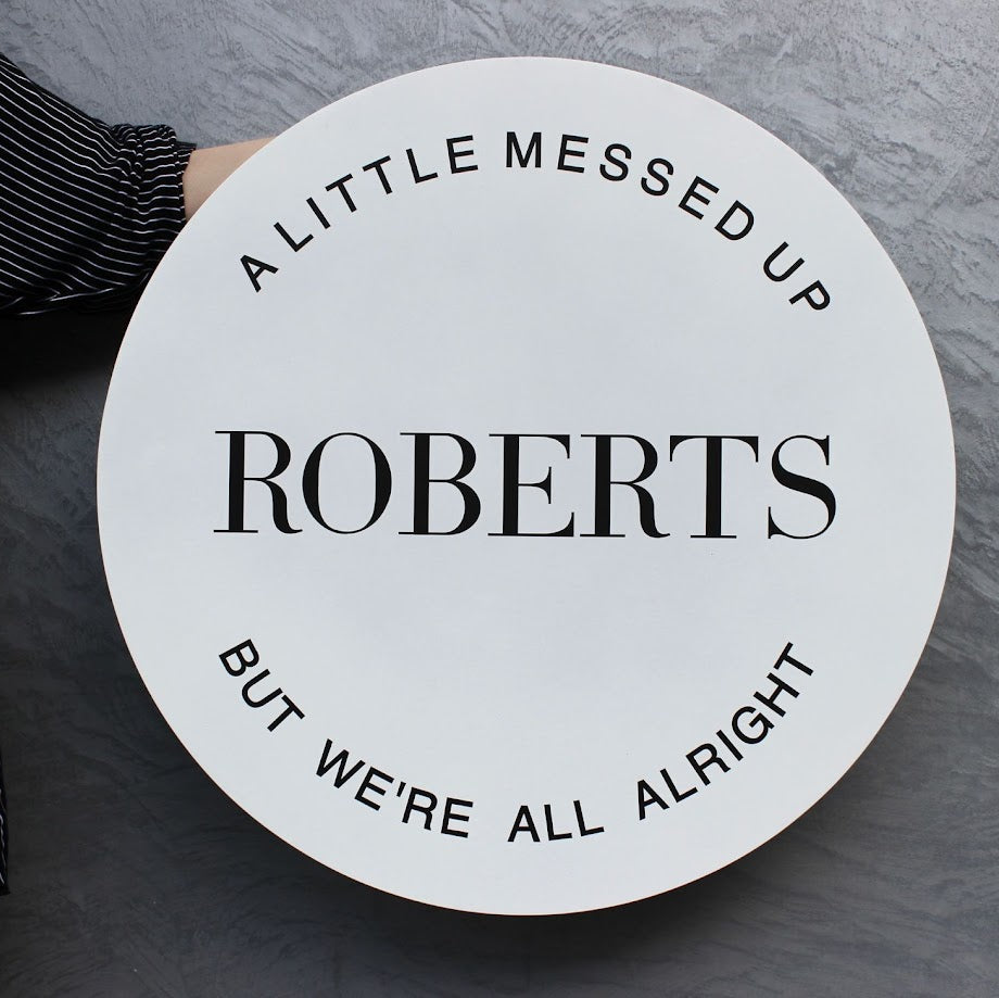 Round sign with text 'A LITTLE MESSED UP ROBERTS BUT WE'RE ALL ALRIGHT' held by a person on a gray background