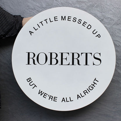 Round sign with text 'A LITTLE MESSED UP ROBERTS BUT WE'RE ALL ALRIGHT' held by a person on a gray background