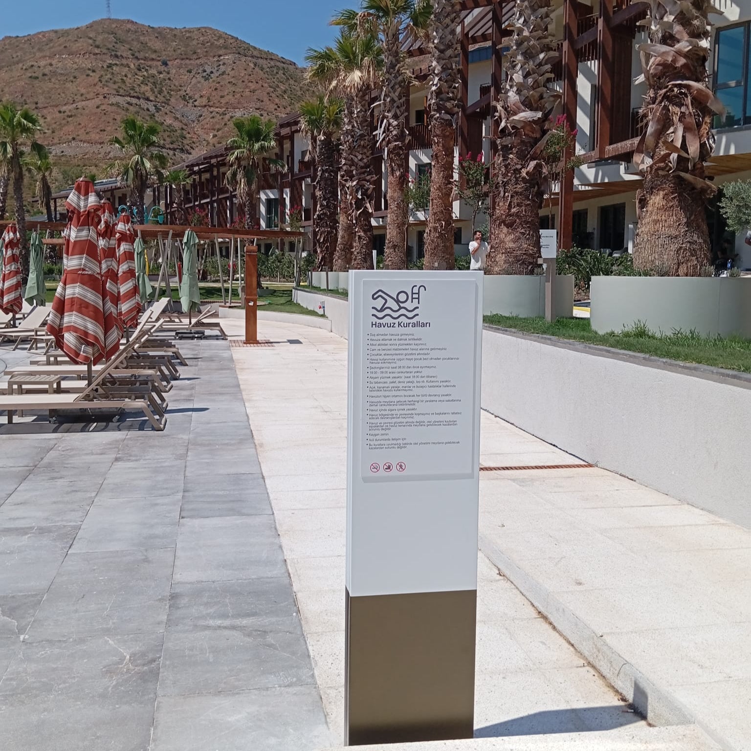 Beachside area with lounge chairs, palm trees, and a sign in front of a building.