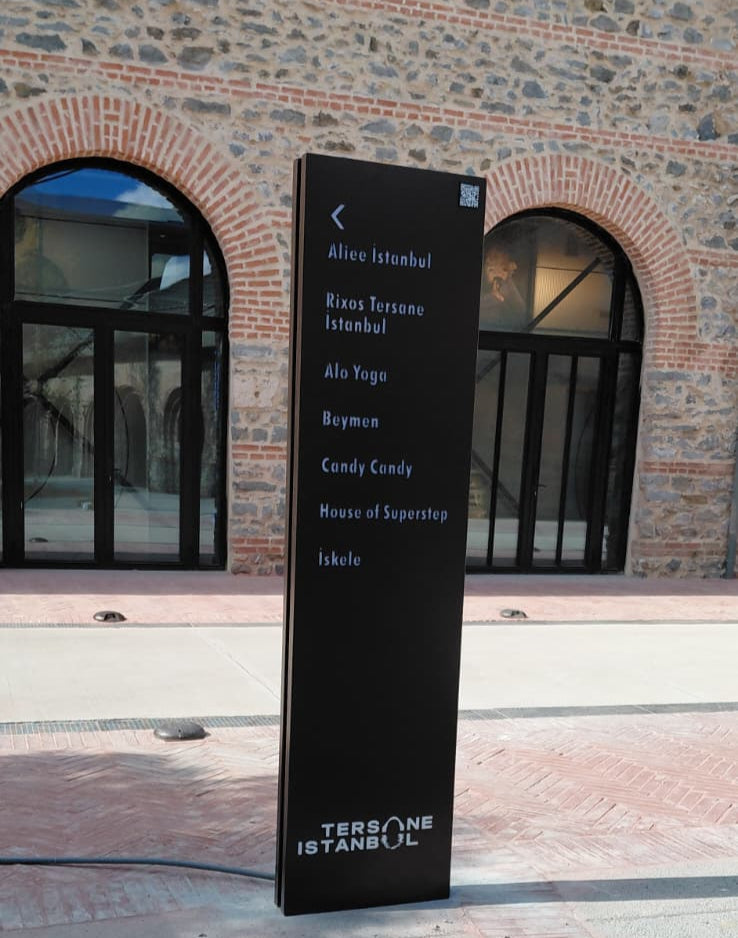 Black vertical sign with text in front of a building with arched windows.