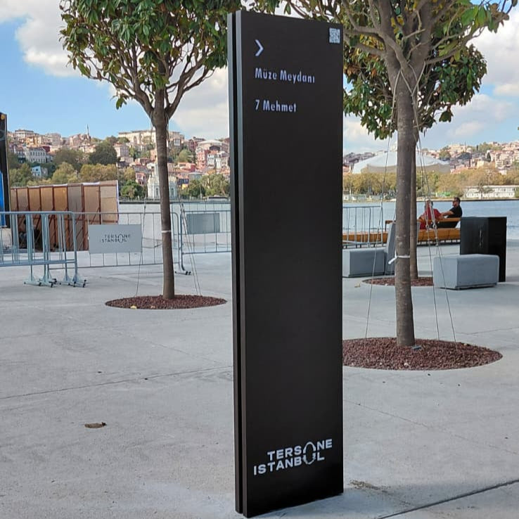 Tall black signpost on a sidewalk with trees and a cloudy sky in the background