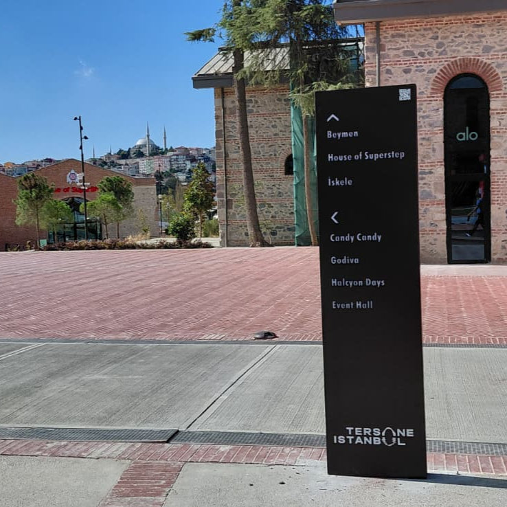 Black directional sign on a sidewalk with a red building and crane in the background