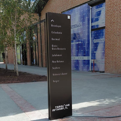 Black directional sign in front of a brick building with large glass windows.
