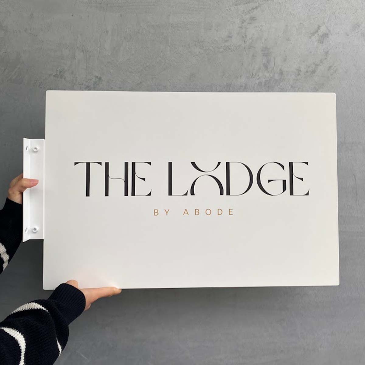 Person holding a large white board with 'The Lodge by Abode' text on a gray background