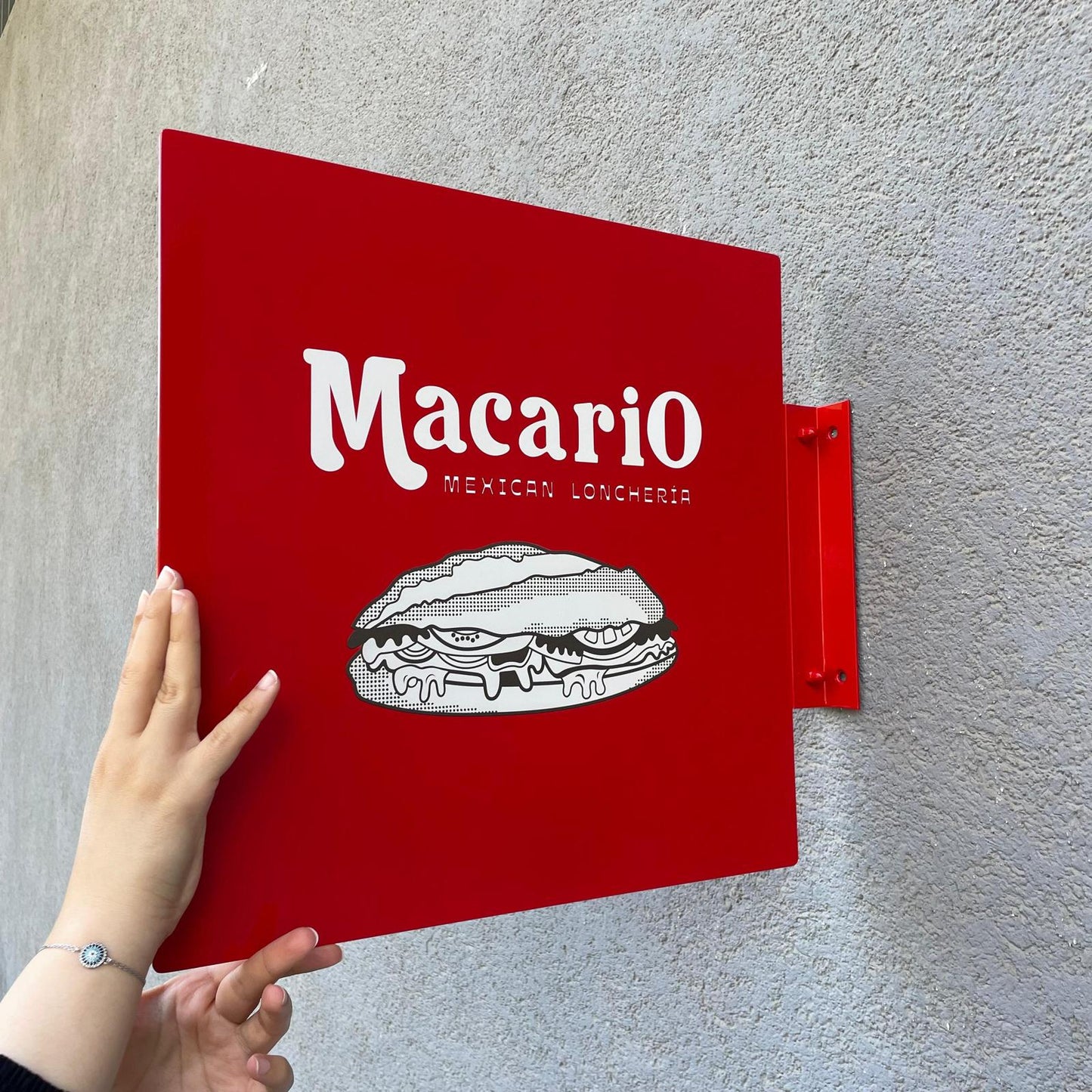 Red sign with 'Macario' and a sandwich illustration on a gray wall.