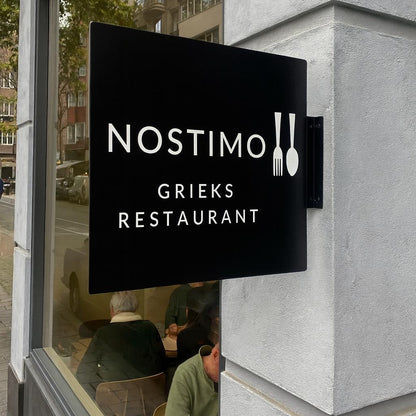 Nostimo Grieks Restaurant sign on a building exterior with reflection of street scene.