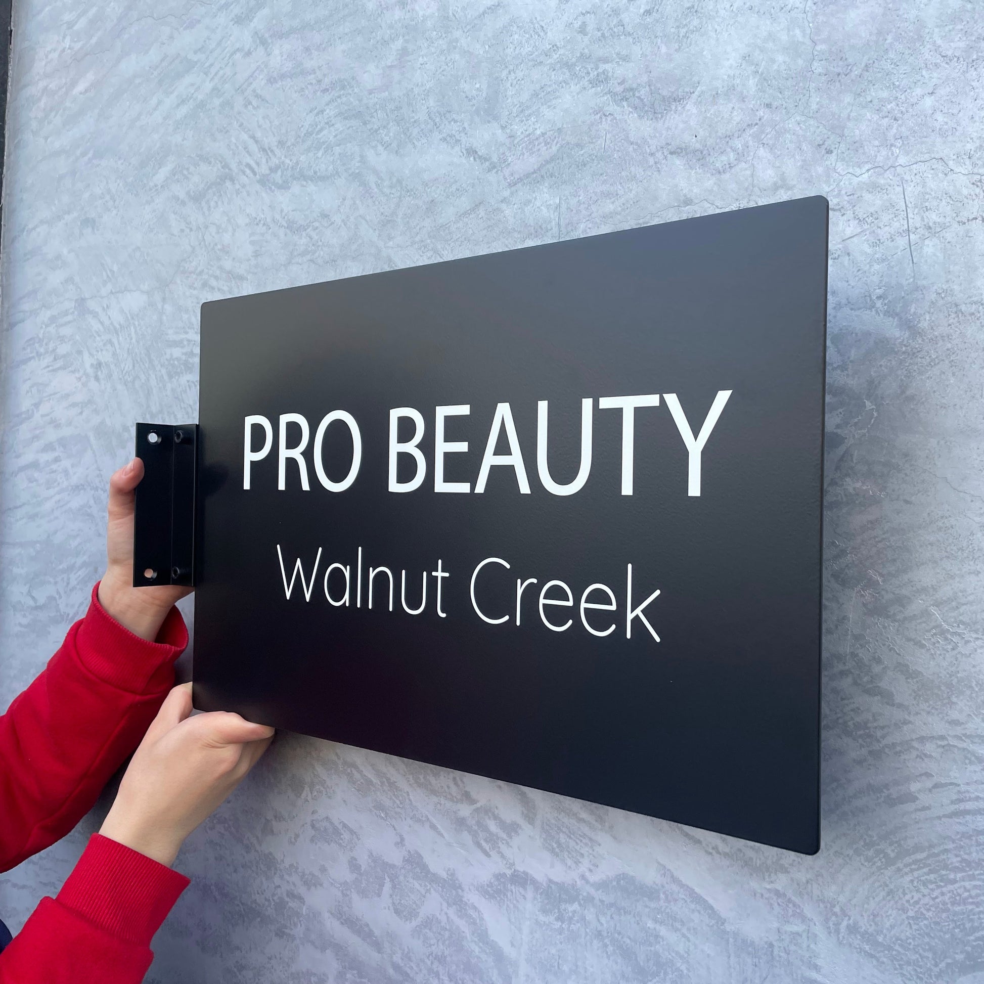 Person holding a black sign with 'PRO BEAUTY Walnut Creek' text against a textured gray wall.