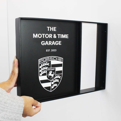 Person holding a black framed sign with Porsche logo and text on a white background