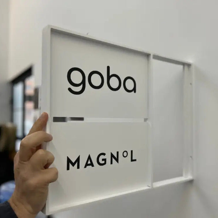 Person holding a frame with 'goba' and 'MAGNOL' branding against a white wall.