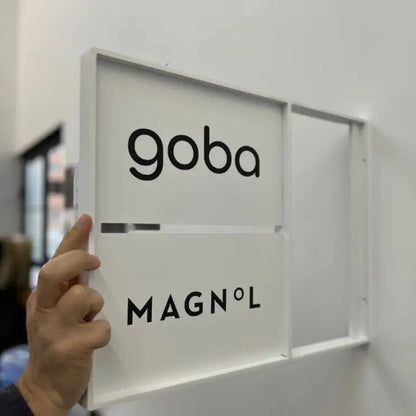 Person holding a frame with 'goba' and 'MAGNOL' branding against a white wall.