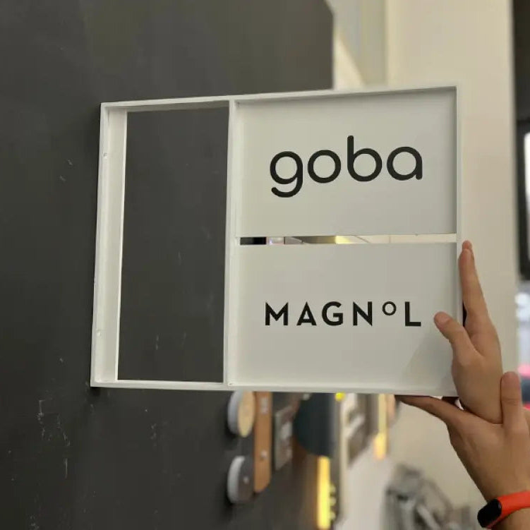 Person holding a frame with 'goba' and 'MAGNÓL' branding against a blurred indoor background.