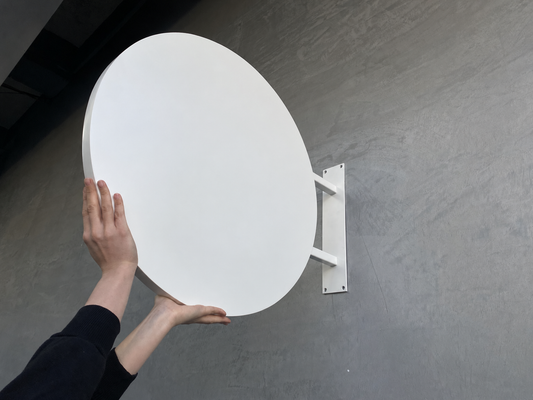 Unprinted white thick round blade sign held for scale, showing two support arms and wall mounting plate; exact diameter not specified